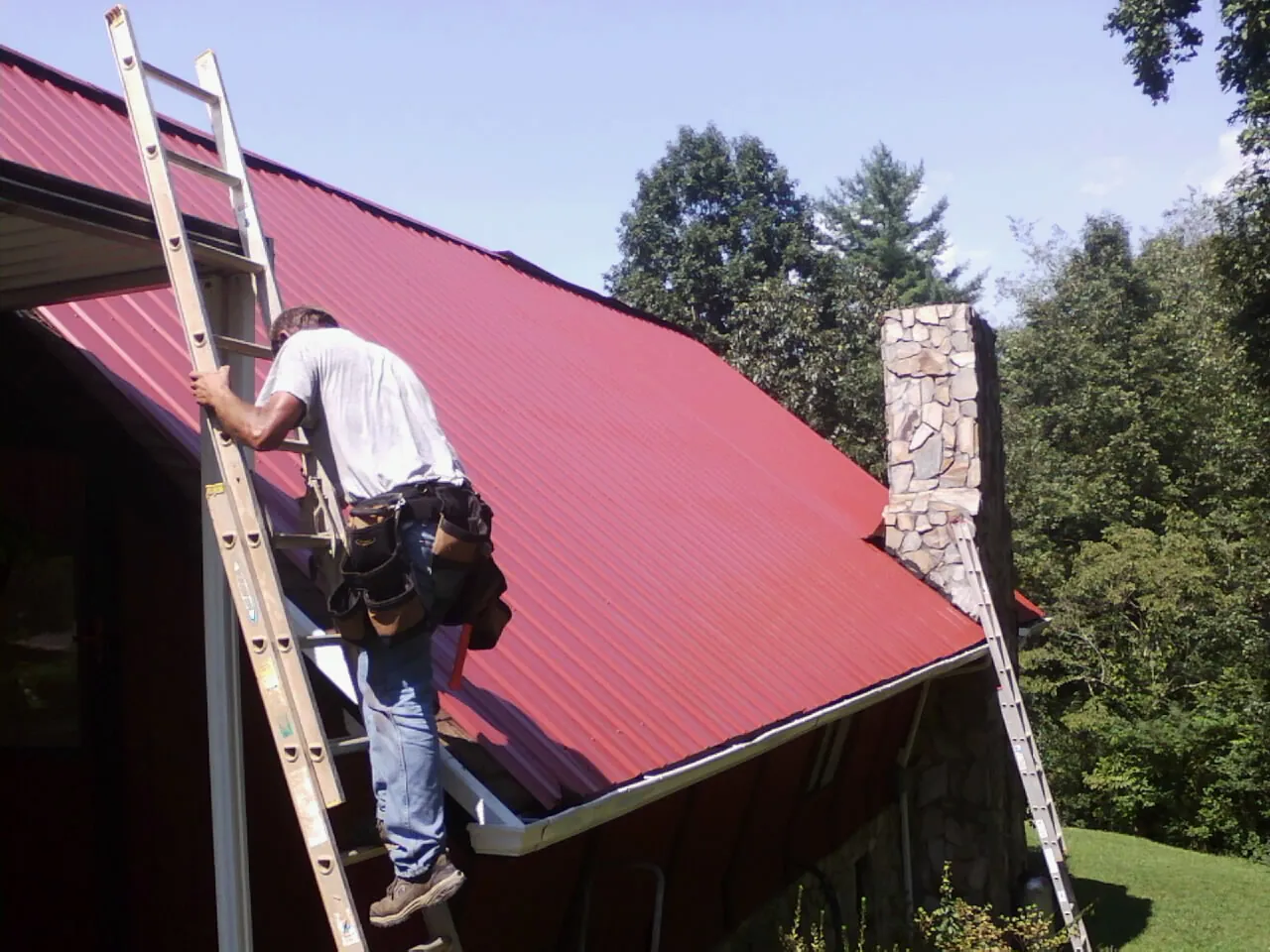 Roofer on a ladder beside a red metal roof and stone chimney — Soffit & Fascia Repair by Roofing Brothers in Rockton